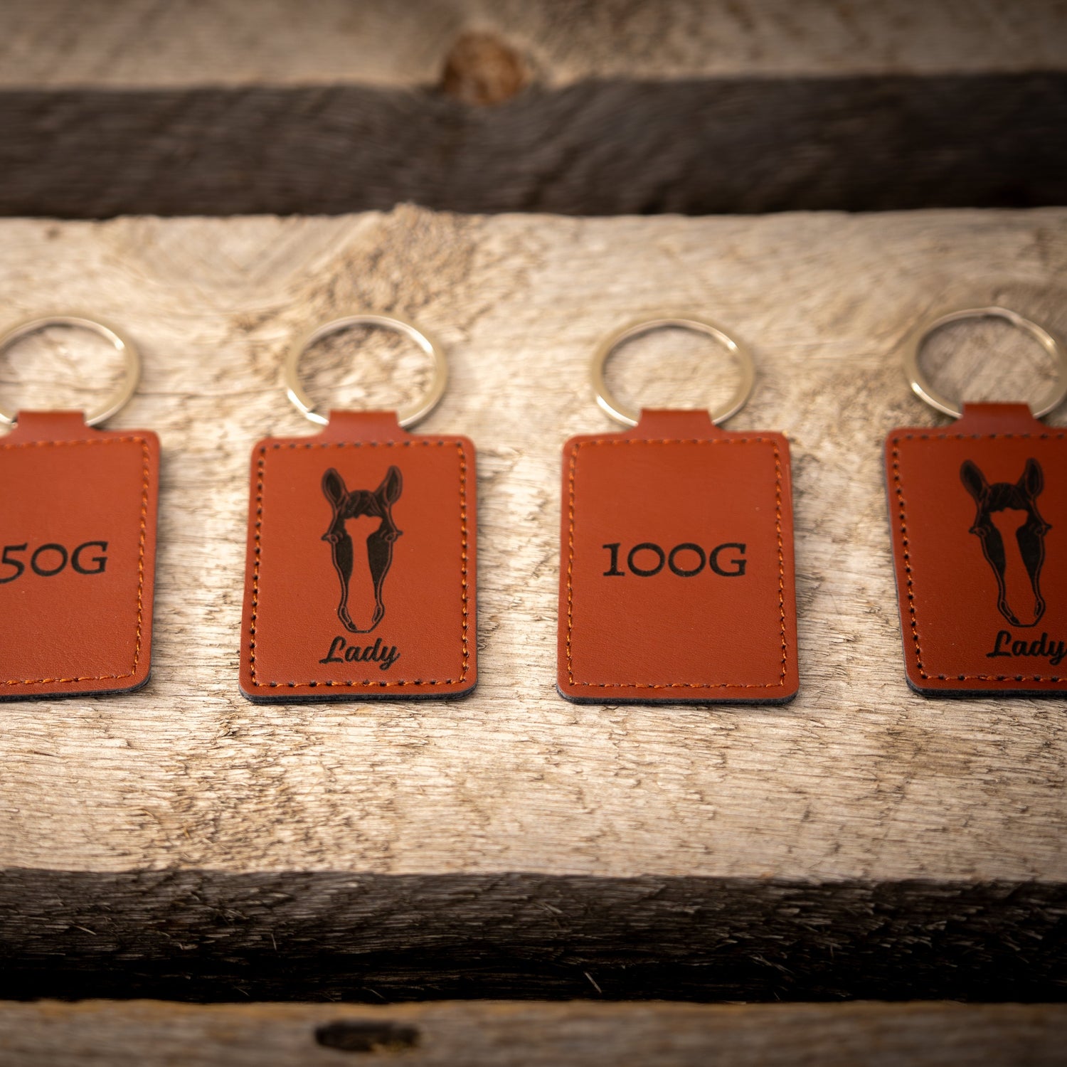 Five brown leather keychains with black horse illustrations and weight labels on a wooden surface.